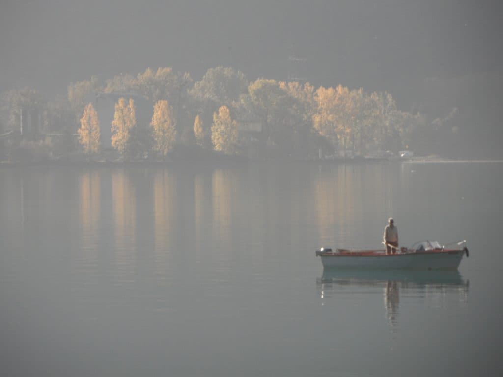 Fisherman in a boat on a lake, with reflections of trees and the man fishing.