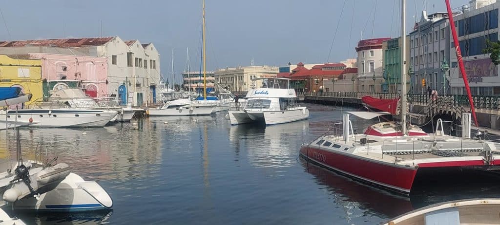 The Sandals dive boat is seen entering the harbor of Port of Bridgetown, Barbados, amidst a picturesque scene of moored yachts and colorful waterfront buildings.