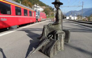 Sculpture of a woman passenger with a wide-brimmed hat, luggage, and an umbrella at a Swiss train station.