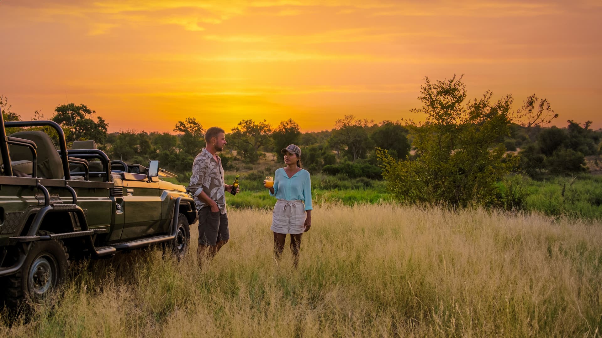 Couple enjoying sundowners in shorts and t-shirts on an African safari with a Land Cruiser, savannah, and trees at sunset