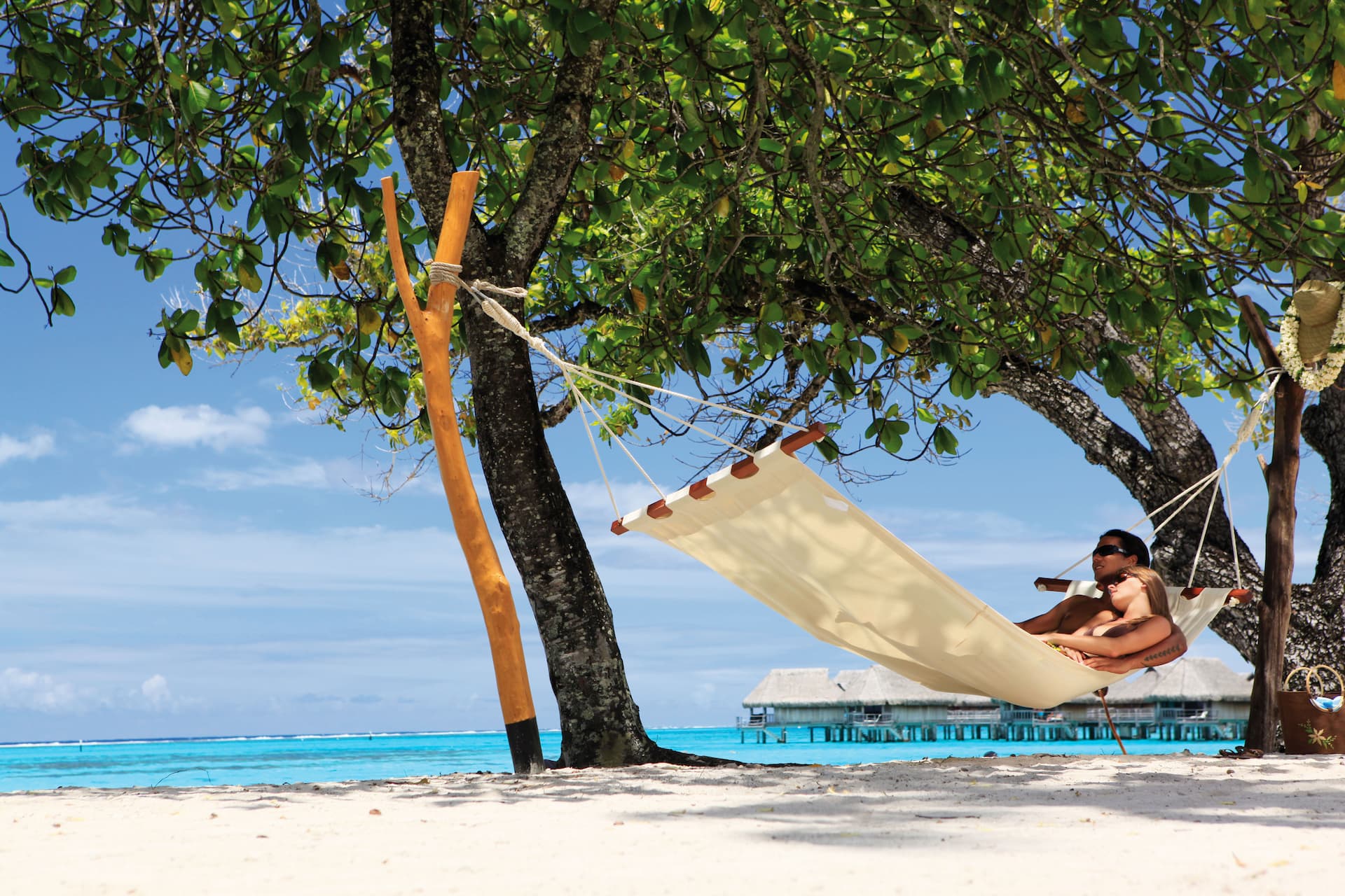 Couple relaxing in a hammock under green trees with Tahitian overwater bungalows in the background over turquoise waters.