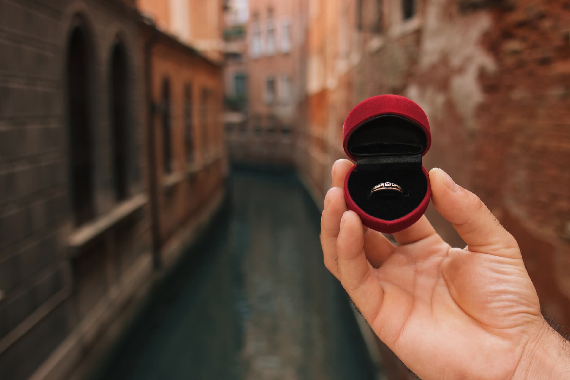 Proposal Trips: A man's hand holding an engagement ring in a red velvet box with a side canal in Venice in the background