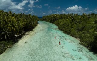 Aerial view of two paddleboarders on a wide channel in Tetiaroa Atoll with shallow, crystal clear water, coral, and fish swimming beneath.