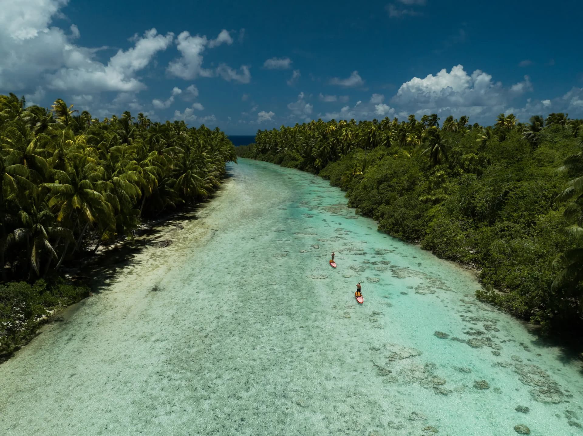 Aerial view of two paddleboarders on a wide channel in Tetiaroa Atoll with shallow, crystal clear water, coral, and fish swimming beneath.