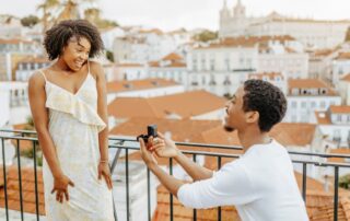 Smiling young African American man kneeling and proposing with a ring to a woman in a dress in a European city