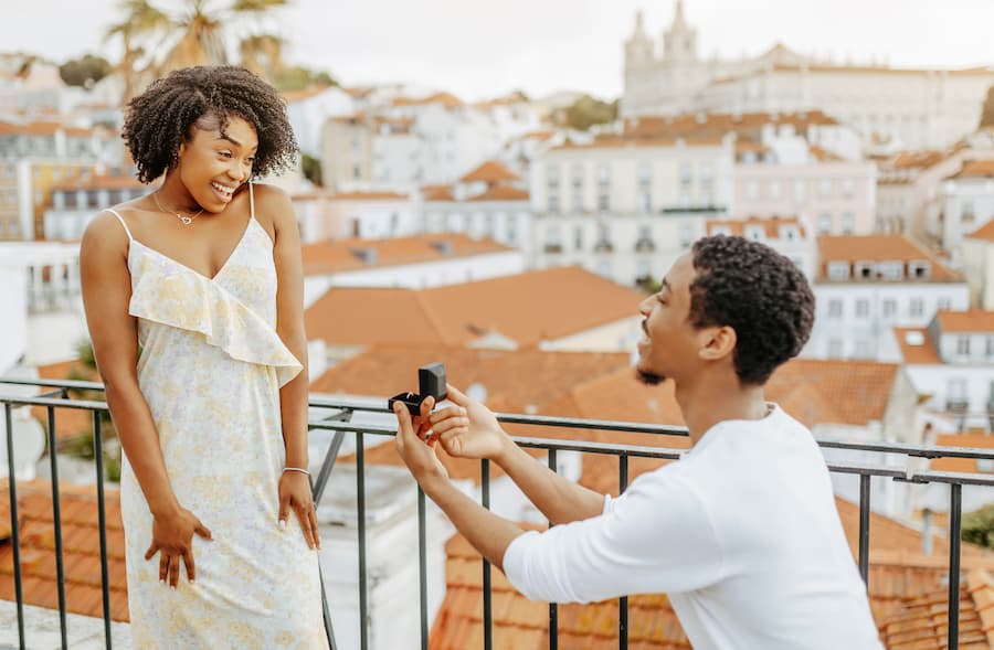 Smiling young African American man kneeling and proposing with a ring to a woman in a dress in a European city