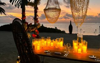 Romantic dinner setup under the stars on the beach with candles casting an orange glow.