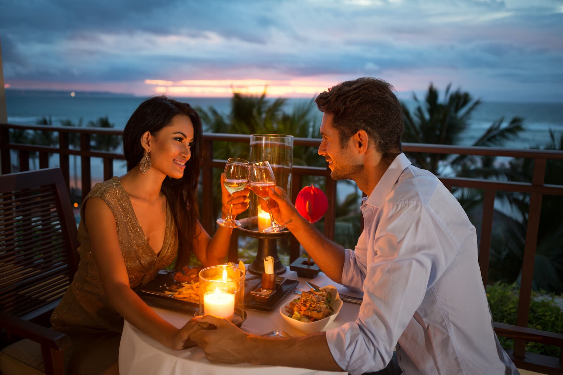 Couple sharing a meal and clicking their wine glasses together with a sunset in the background