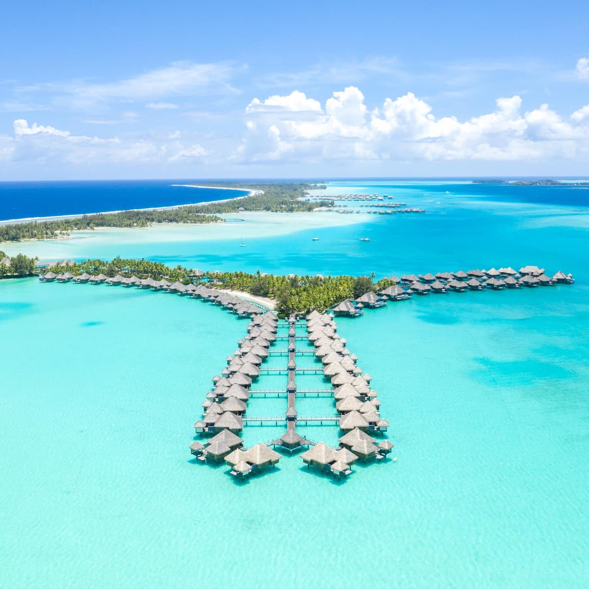 Overwater bungalows of The St. Regis Bora Bora Resort from overhead with crystal clear water in the lagoon and darker Pacific water outside.