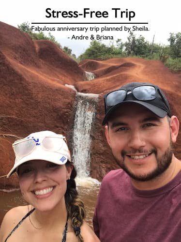 A happy couple on their stress-free anniversary trip to Kauai, smiling with the Red Dirt Waterfall behind them, planned by Sheila Cannon.